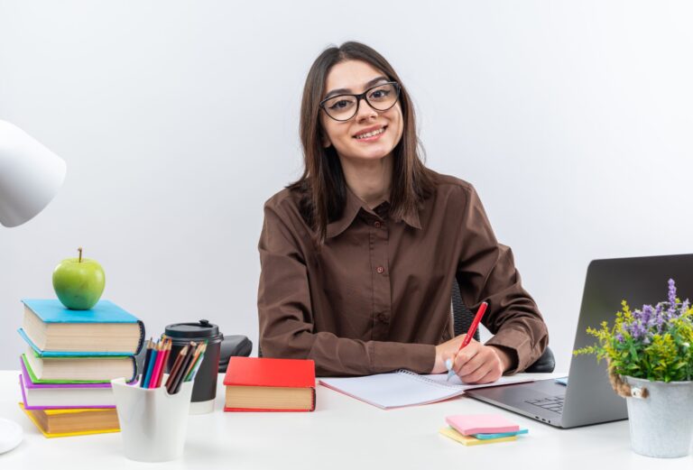 smiling young school girl wearing glasses sits at table with school tools writing something on notebook isolated on white background